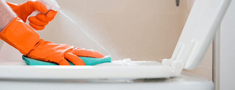 Mãos usando luvas de borracha laranja borrifam produto de limpeza em spray e passam um pano verde sobre a tampa levantada de um vaso sanitário branco.