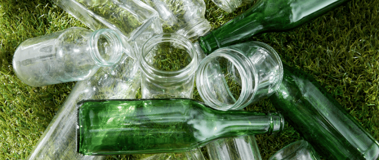 Várias garrafas e potes de vidro vazios, nas cores verde e transparente, espalhados sobre um gramado verde.