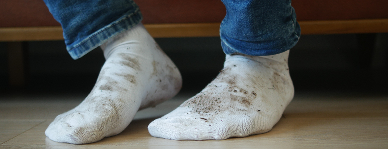 Pés de uma pessoa usando meias brancas curtas com manchas de terra e sujeira, apoiados sobre piso de madeira clara.
