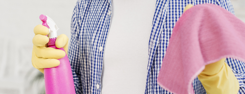 Pessoa com camiseta branca e camisa azul, usando luvas amarelas segura um borrifador rosa e um pano microfibra para limpeza.