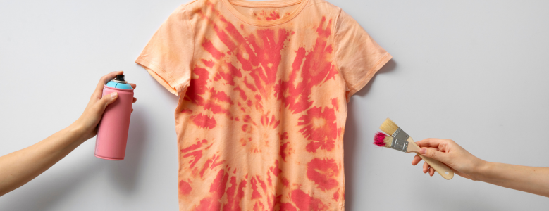 Camiseta estilo tie-dye em tons de laranja e vermelho estendida em um fundo claro, com duas mãos segurando ferramentas de pintura (uma lata de spray rosa e pincéis) nas laterais.
