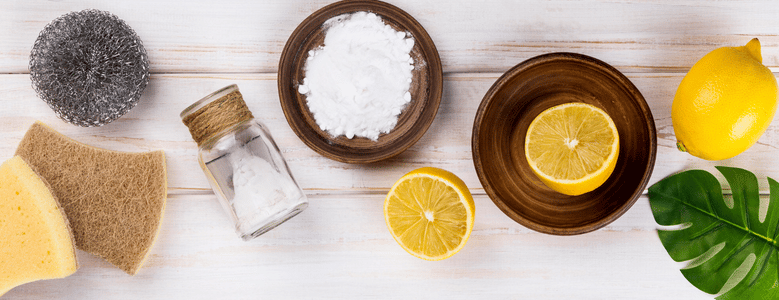 Vista de cima de produtos de limpeza natural sobre uma mesa de madeira branca: esponja de aço, esponjas amarelas, um frasco de vidro com bicarbonato de sódio, uma tigela de madeira com bicarbonato de sódio, limões inteiros e cortados em tigelas de madeira e uma folha verde de costela-de-adão.