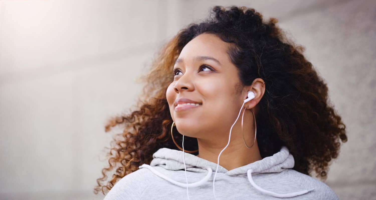 Mulher jovem de cabelo cacheado volumoso, usando moletom cinza com capuz e fones de ouvido intra-auriculares brancos com fio. Ela olha para cima com um leve sorriso, em frente a uma parede clara.