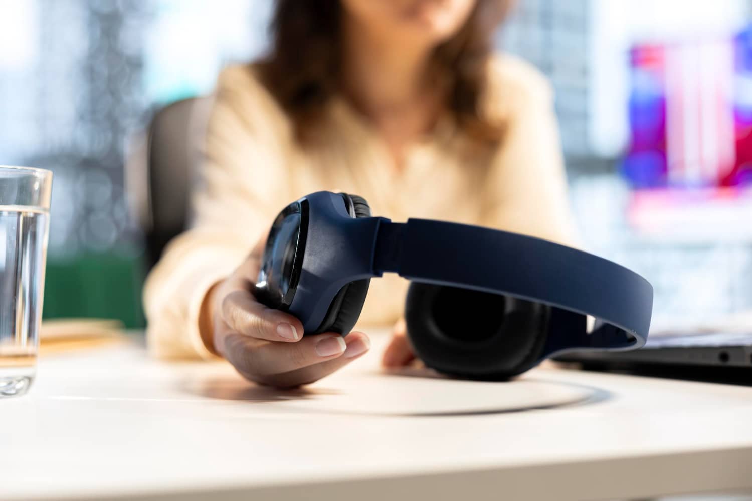 Mulher sentada a uma mesa branca de escritório, com o rosto desfocado ao fundo, apoiando um fone de ouvido azul-marinho over-ear sobre a mesa. À esquerda, um copo de água; ao fundo, telas e ambiente de trabalho desfocados.