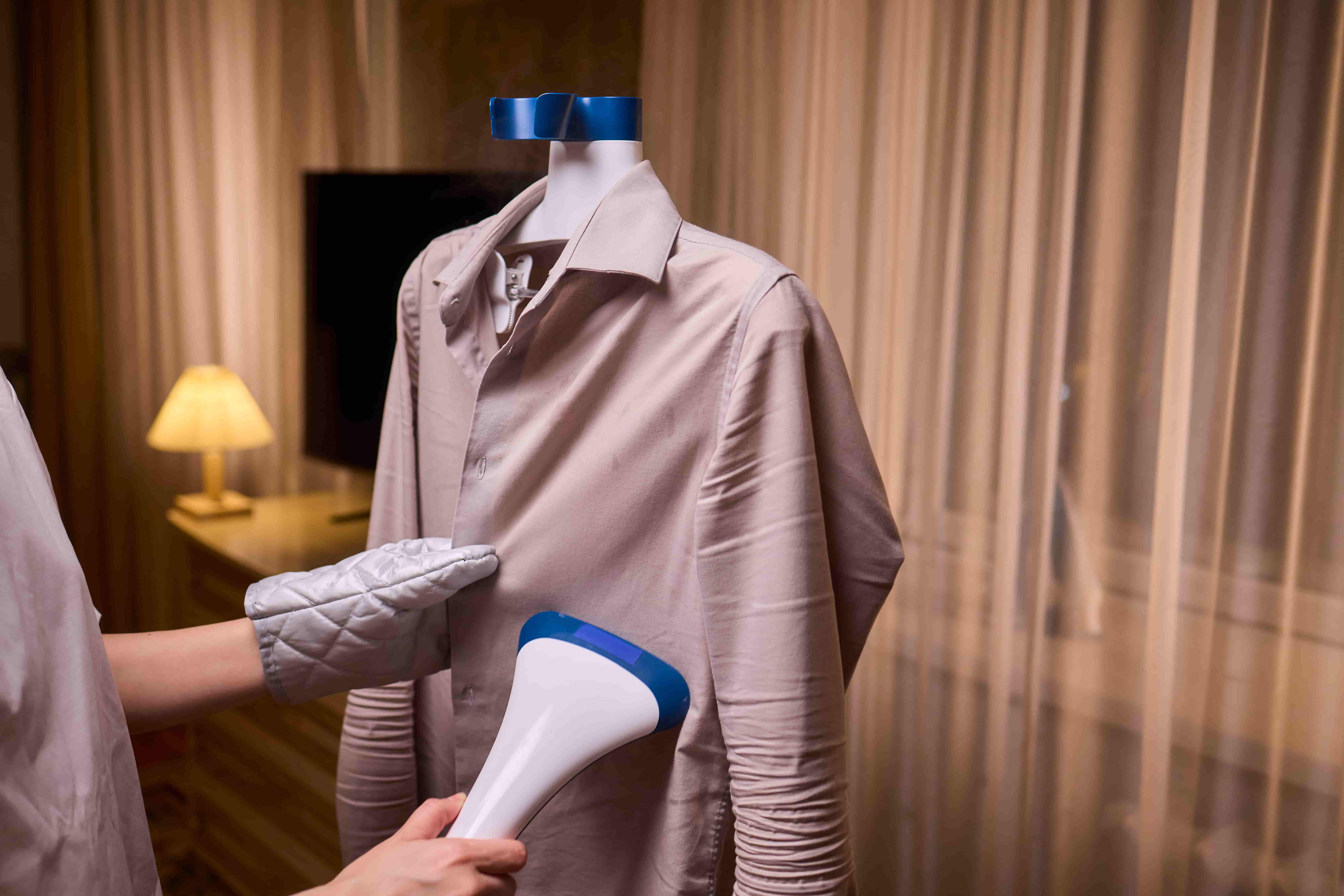 Pessoa usando luva térmica e passando vaporizador em camisa bege pendurada em manequim, em ambiente com iluminação quente.
