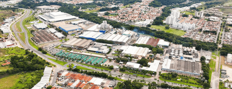 Vista aérea de um grande complexo industrial composto por diversos galpões, estruturas fabris e tanques de armazenamento. A fábrica é cercada por extensas áreas arborizadas, bairros residenciais ao fundo e uma rodovia com curvas no lado esquerdo.