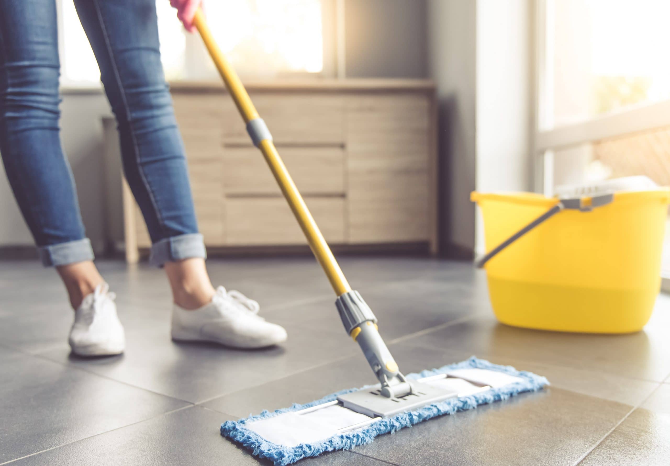 Pessoa de cal&ccedil;a jeans e t&ecirc;nis brancos limpando piso de porcelanato cinza com mop de cabo amarelo e refil azul. Ao lado, um balde amarelo, com uma c&ocirc;moda de madeira ao fundo e luz do sol entrando pela janela.
