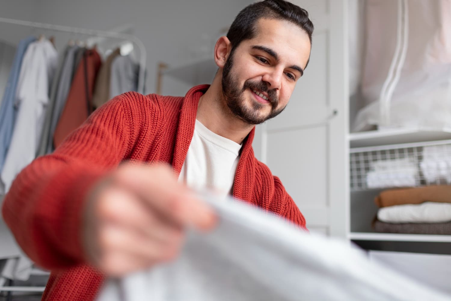 Homem com barba, sorrindo e vestindo um casaco vermelho, dobrando uma peça de roupa branca. Ao fundo, um guarda-roupa branco e organizado, com roupas penduradas e dobradas nas prateleiras.