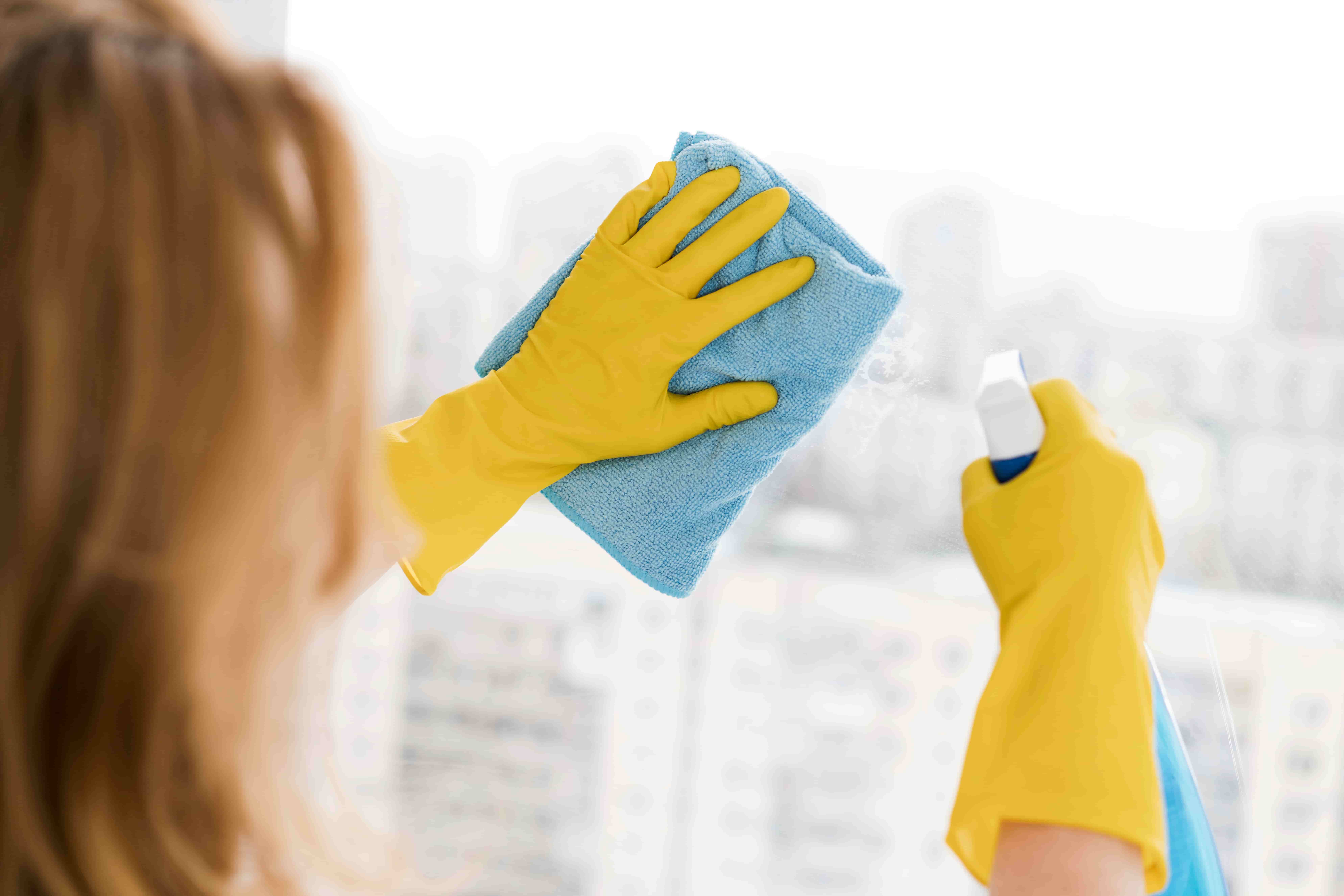 Mulher com luvas amarelas limpando janela de vidro com pano de microfibra azul e borrifador.