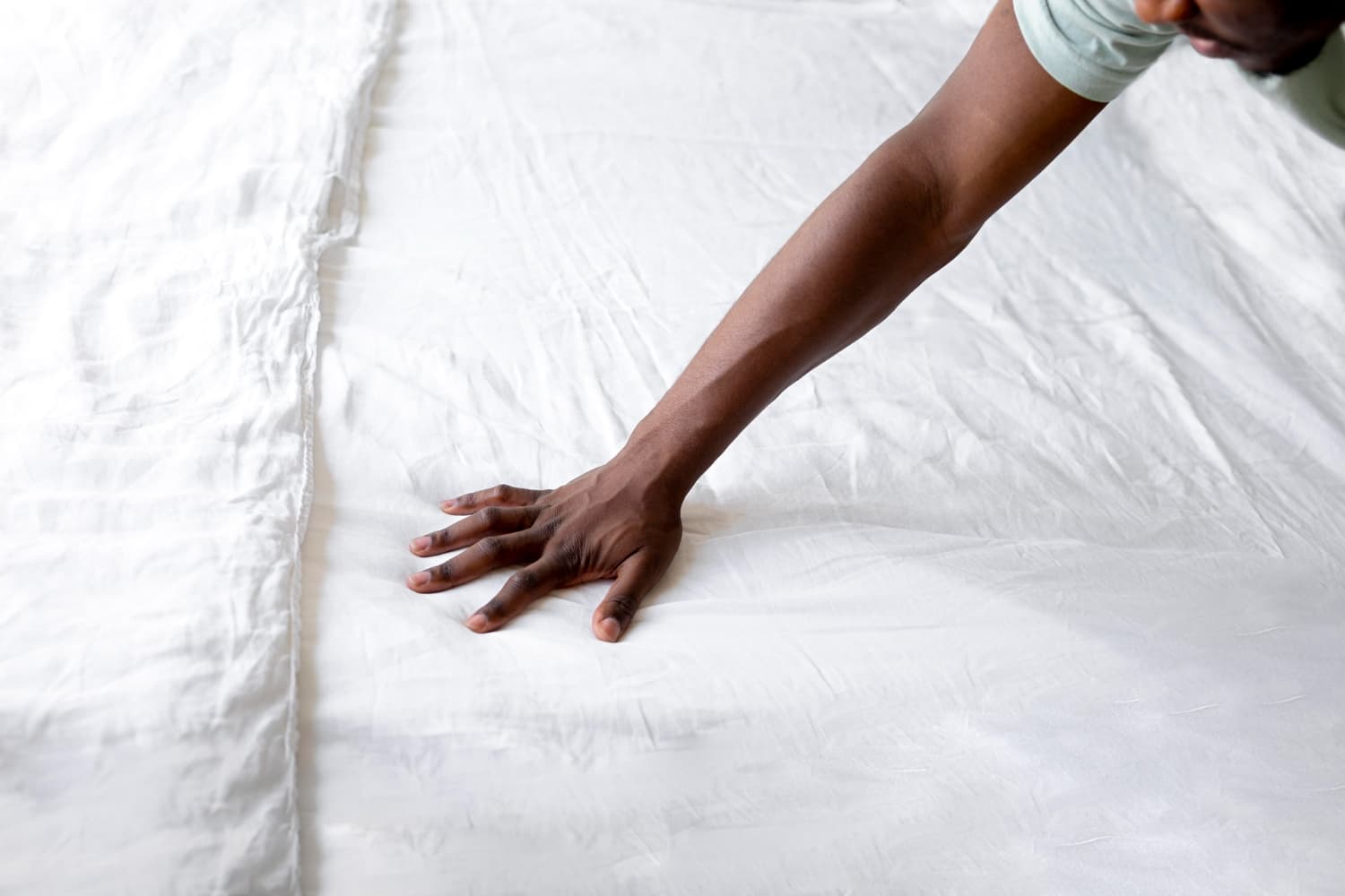 Bra&ccedil;o e m&atilde;o de uma pessoa de pele negra estendida, tocando e alisando um len&ccedil;ol branco sobre uma cama.