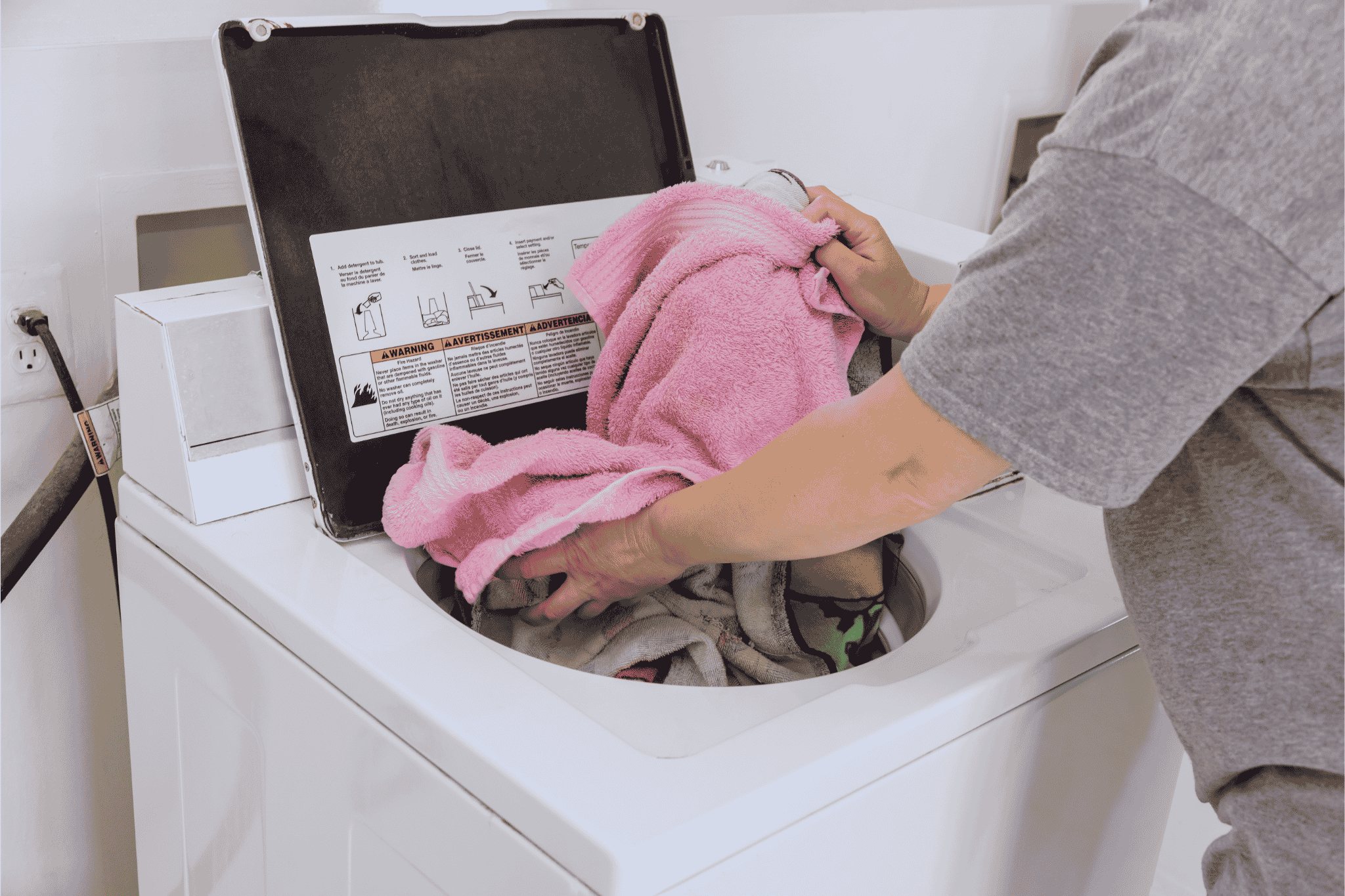 Pessoa colocando uma toalha rosa dentro de uma m&aacute;quina de lavar branca de abertura superior.