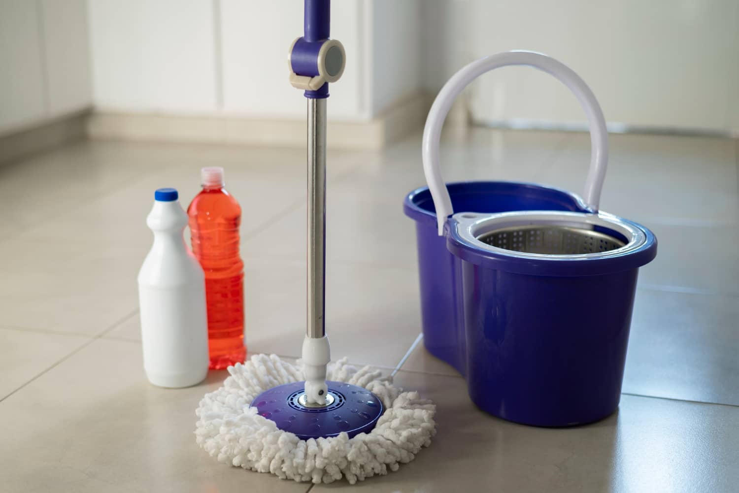 Mop girat&oacute;rio de microfibra branca com cabo azul apoiado em piso claro, ao lado de um balde azul com cesto centrifugador e dois frascos de produtos de limpeza (um branco e um laranja).