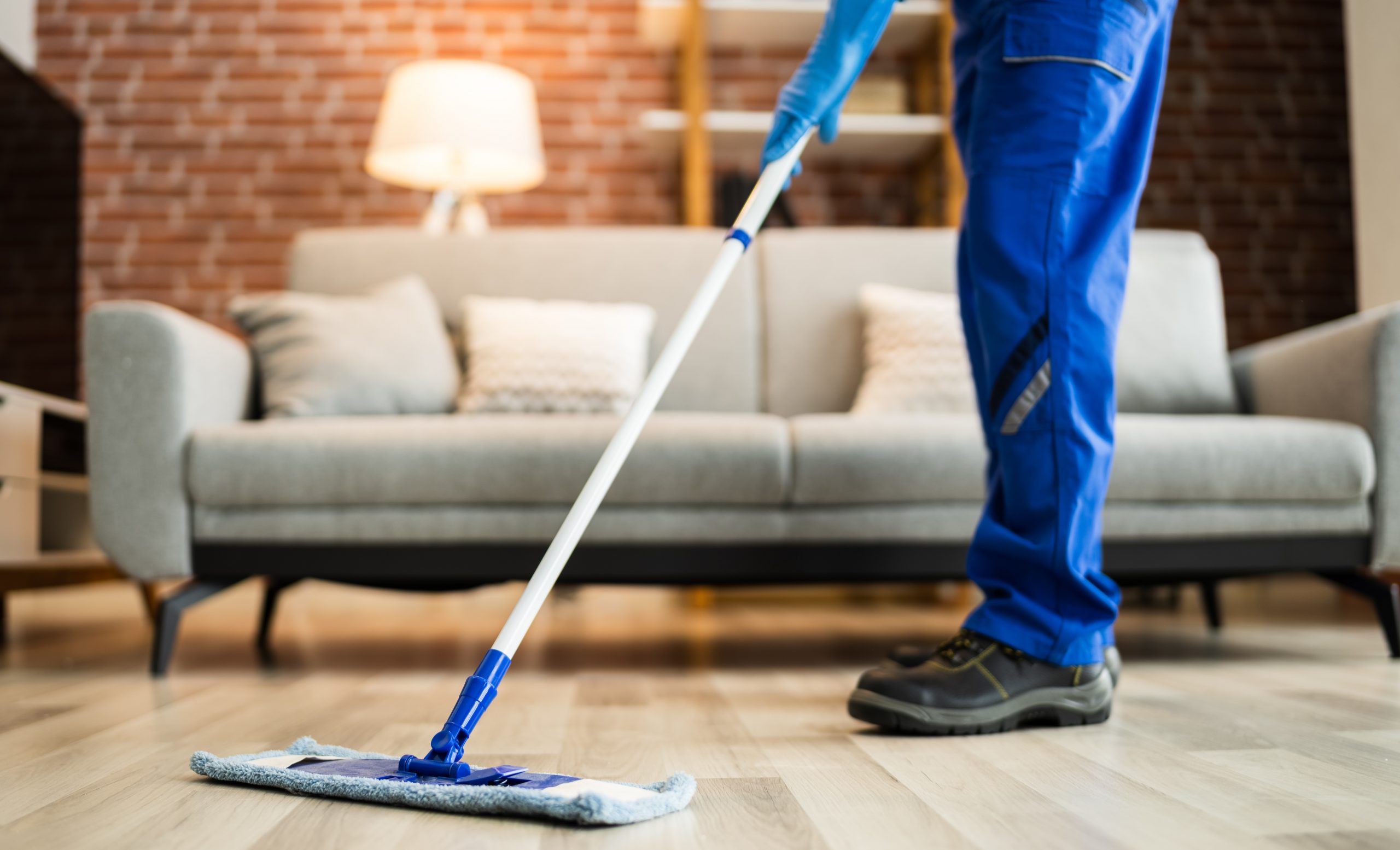Visão parcial das pernas de uma pessoa de calça a azul e sapato fechado preto, passando um mop flat sobre um piso laminado de madeira. Ao fundo observa-se o ambiente de uma sala com sofá e abajur