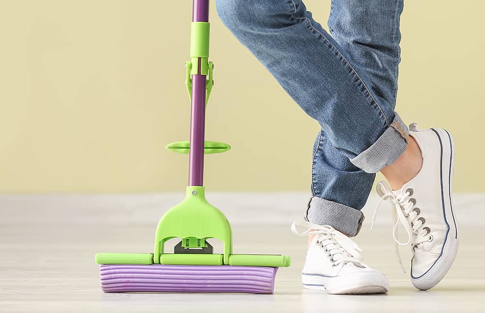 Vista lateral das pernas de uma pessoa de cal&ccedil;a jeans e t&ecirc;nis brancos ao lado de um mop de rolo com esponja lil&aacute;s e estrutura verde, sobre piso claro e fundo amarelo.