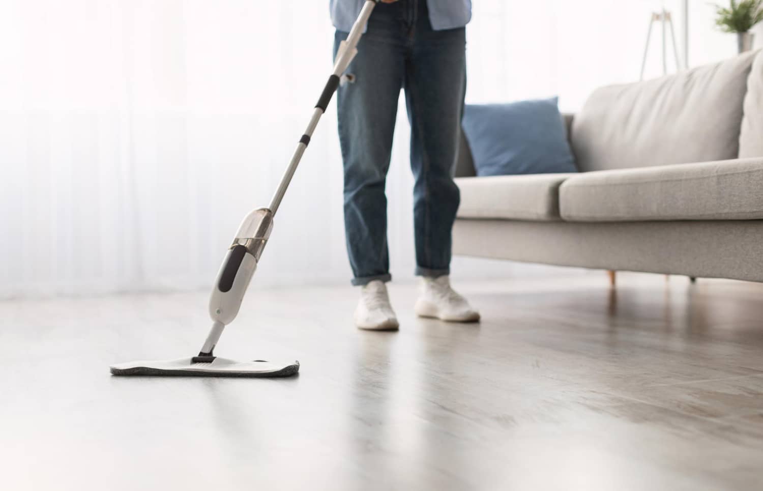 Pessoa de cal&ccedil;a jeans e t&ecirc;nis brancos limpando piso de madeira claro com mop spray de refil de microfibra. Ao fundo, um sof&aacute; cinza com almofada azul em uma sala iluminada.