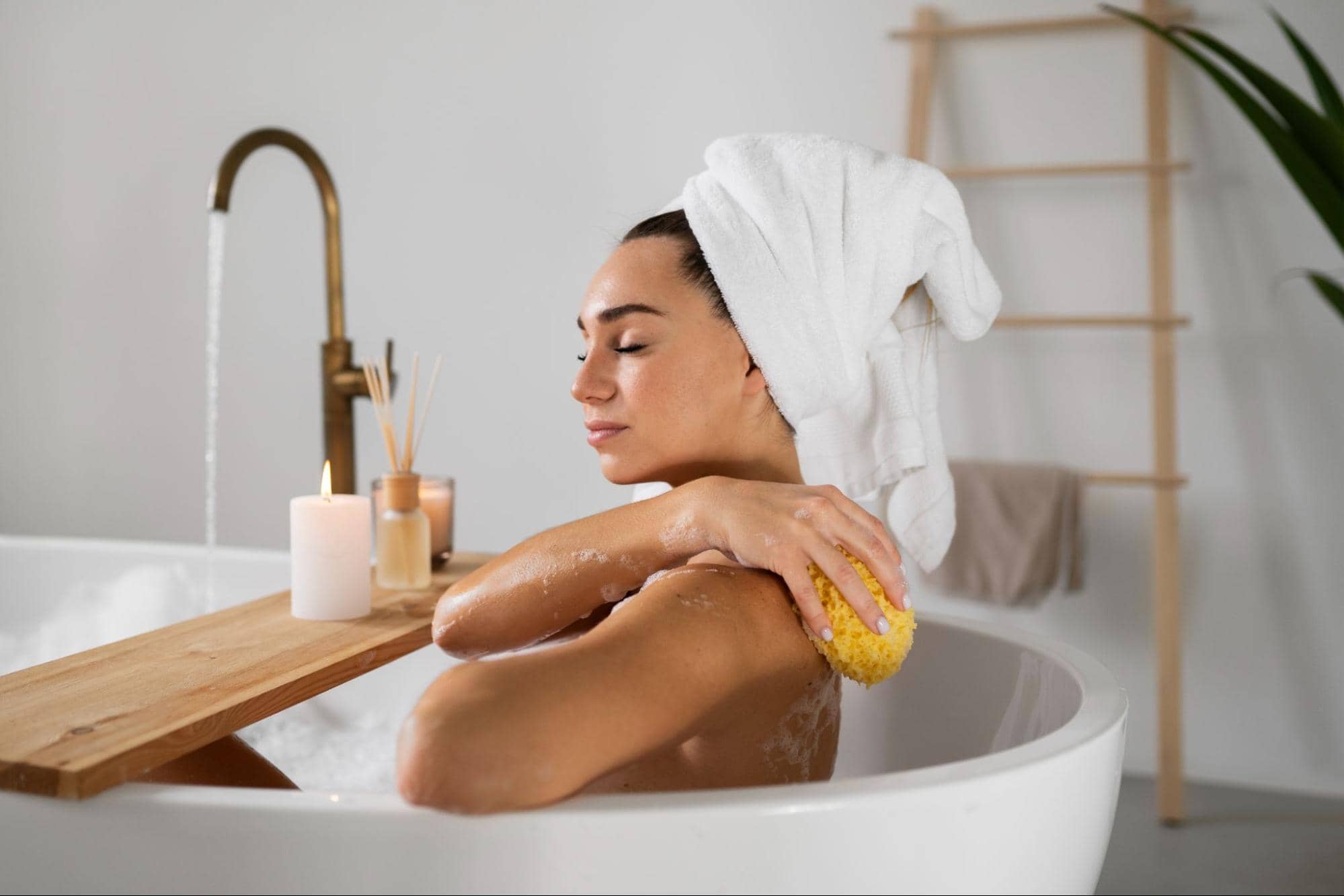 Mulher com toalha na cabe&ccedil;a, tomando banho dentro da banheira com espuma, velas e aromatizantes.