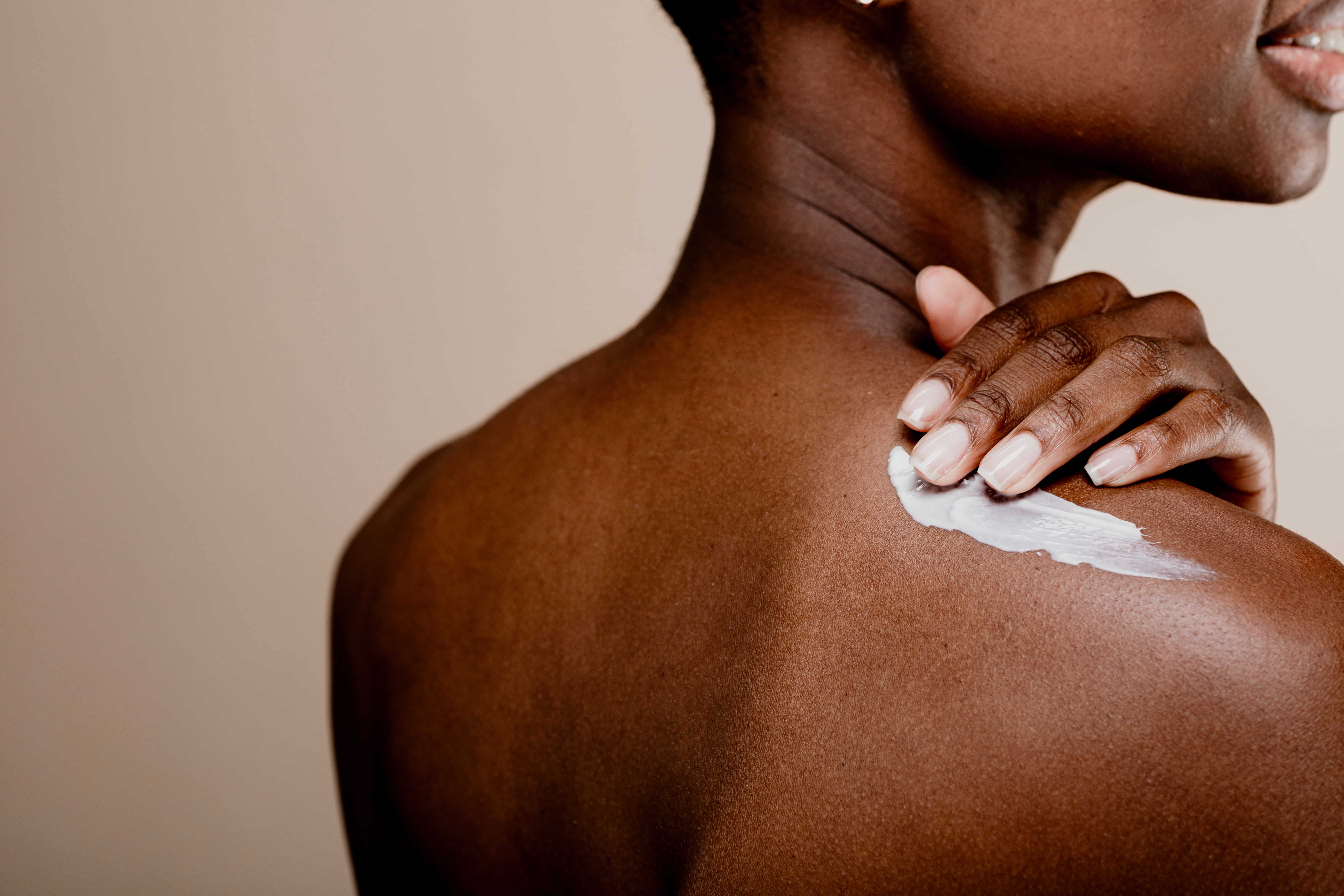 Mulher negra aplicando creme hidratante no ombro e nas costas durante a rotina de bodycare.
