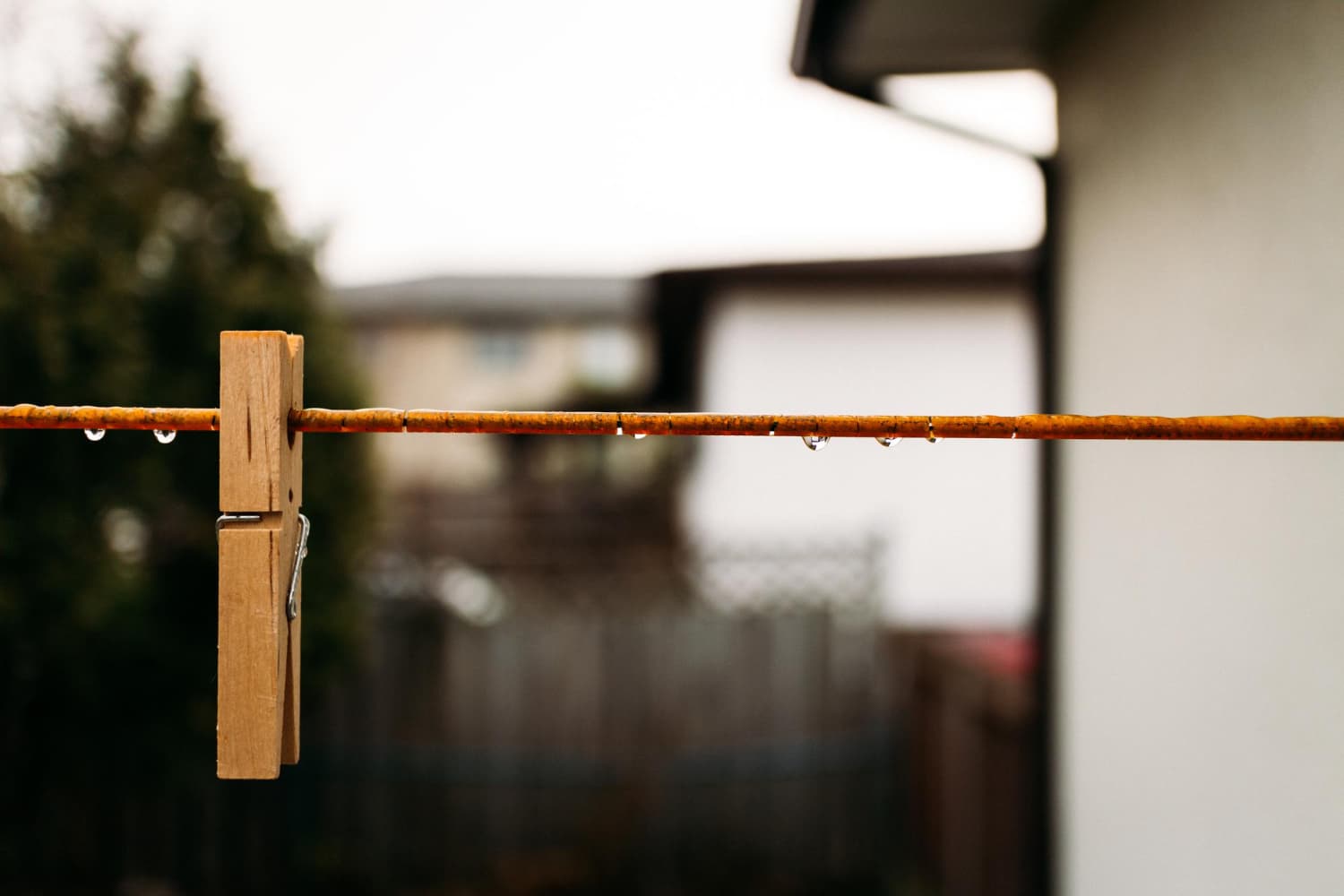 Um prendedor de roupa de madeira preso em um varal laranja com gotas de água. Fundo desfocado mostrando árvores e parte de uma casa.
