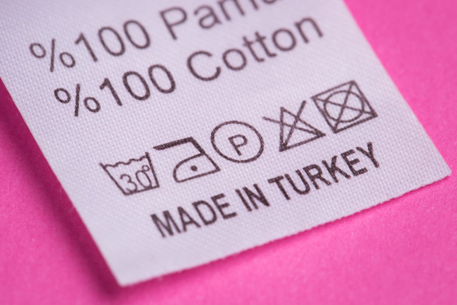 Etiqueta de roupa branca sobre fundo rosa indicando composi&ccedil;&atilde;o &ldquo;%100 Cotton&rdquo;, fabrica&ccedil;&atilde;o &ldquo;MADE IN TURKEY&rdquo; e cinco s&iacute;mbolos de cuidados: lavar a 30&deg;C, passar a baixa temperatura, lavagem a seco, n&atilde;o alvejar e n&atilde;o secar na m&aacute;quina.