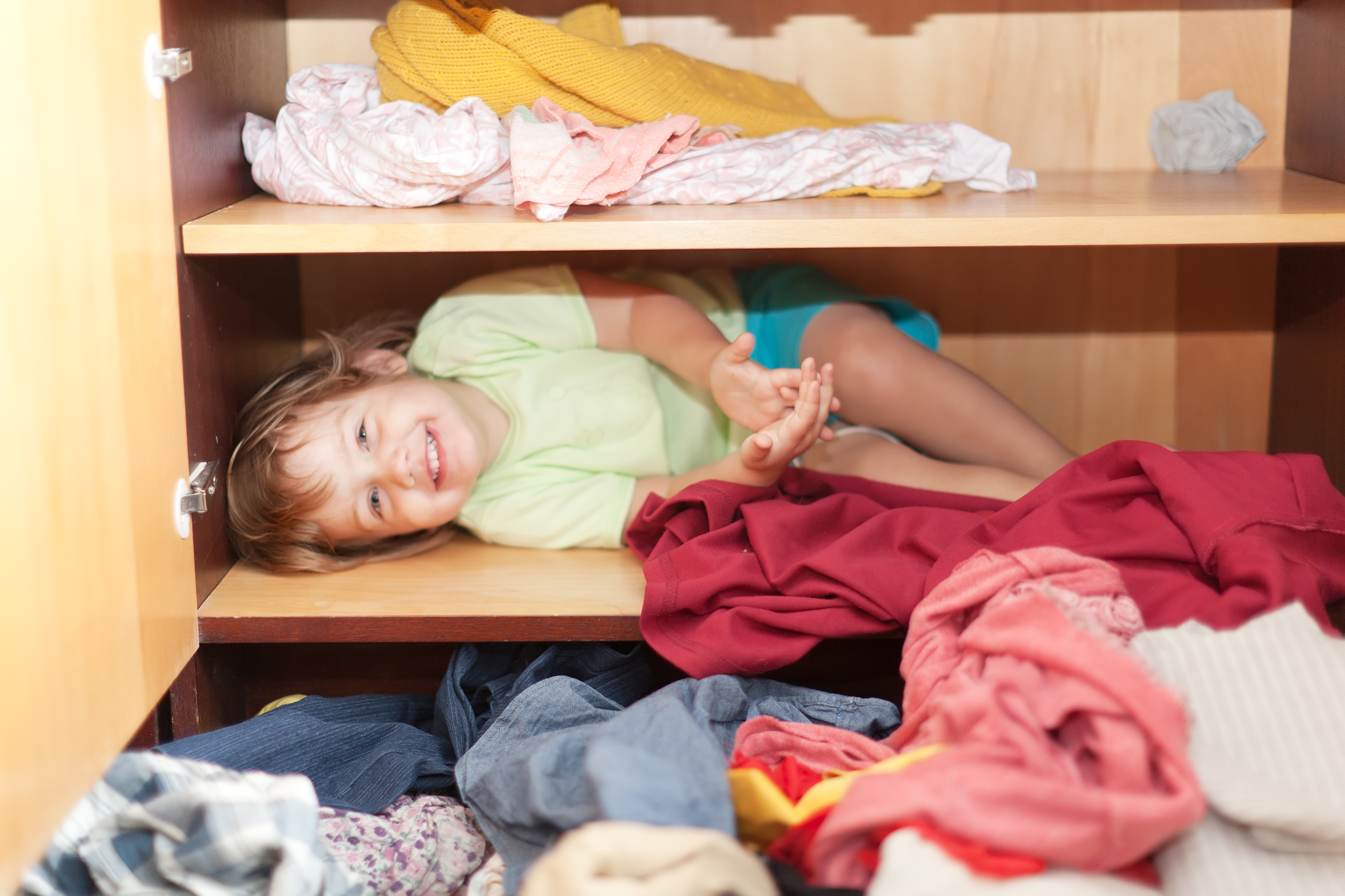Visão aproximada de um guarda-roupa de madeira com a porta e gavetas abertas. No interior do guarda-roupa observa-se várias peças de roupas reviradas e uma criança pequena deitada em uma prateleira sorrindo.