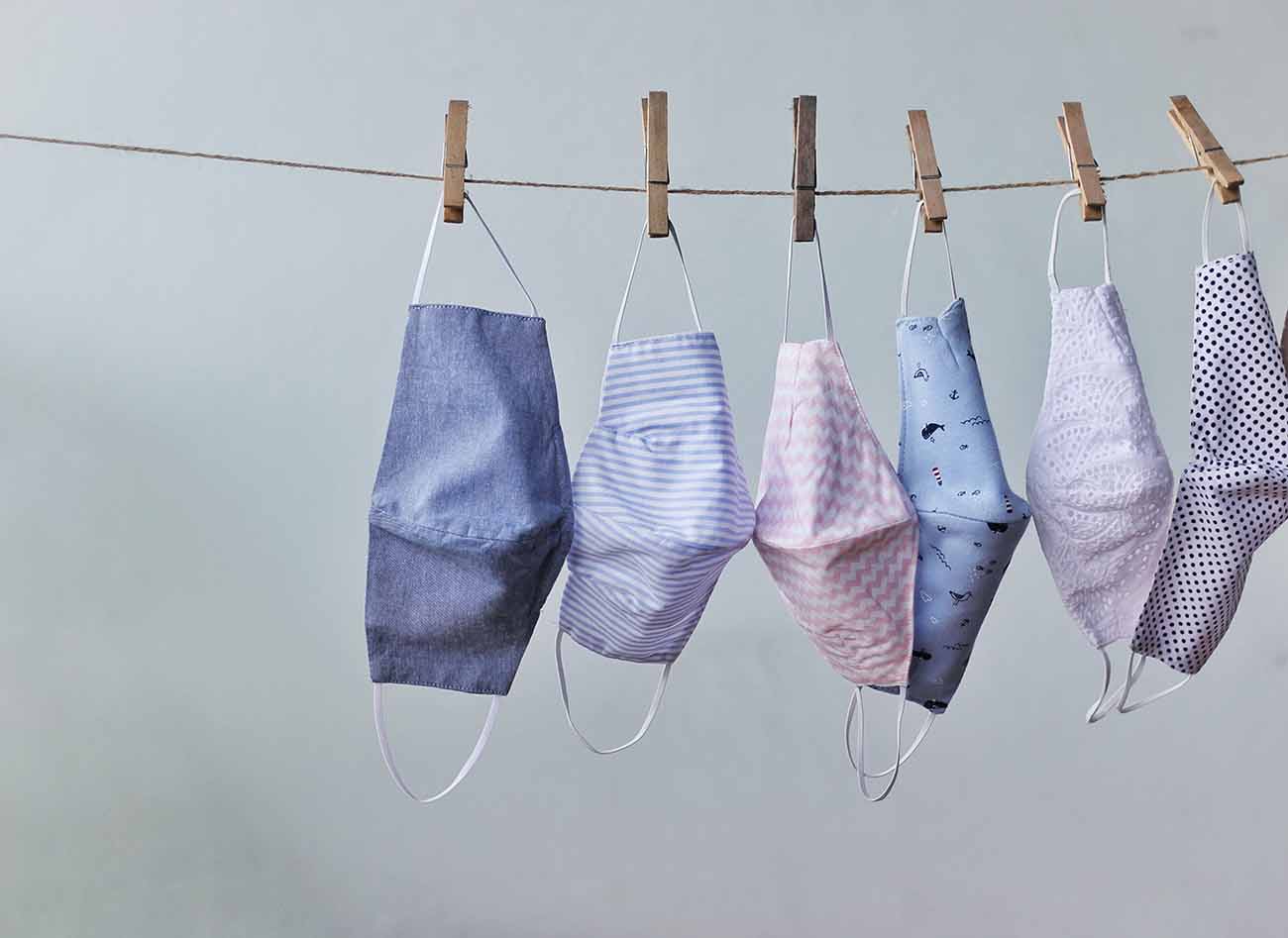 Varal de corda estendido com 6 máscaras faciais de tecido de algodão penduradas para secar na frente de uma parede clara.